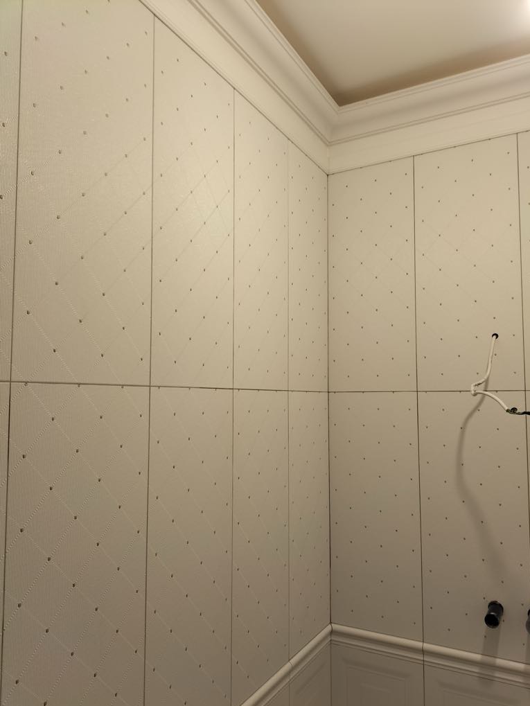 Ściana łazienki wyłożona białymi panelami z delikatnym wzorem rombów i punktów, widoczne łączenia paneli, listwa przypodłogowa i fragment instalacji.
