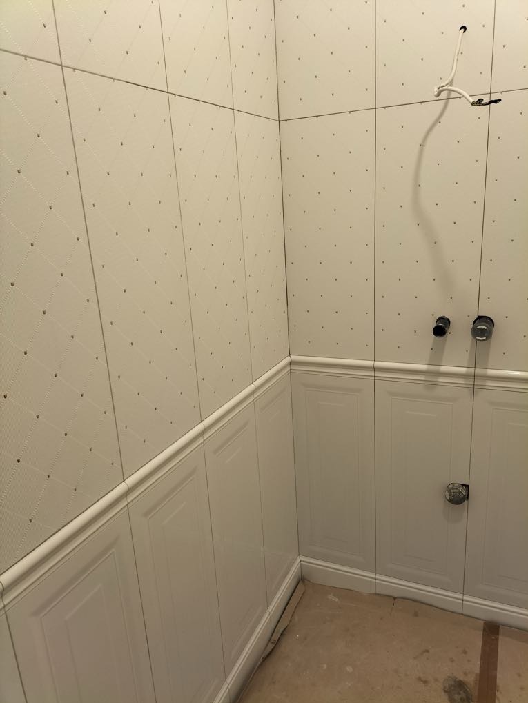 Wykończona łazienka z białymi, wzorzystymi płytkami na ścianach i dolną częścią wykończoną panelami. Widoczne elementy instalacji wodnej.