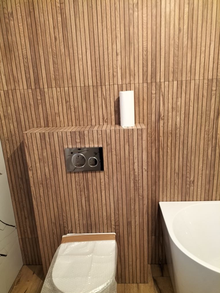 Wykończona łazienka z drewnopodobnymi płytkami na ścianach, częściowo zakryta toaleta i fragment wanny. Widoczny przycisk spłuczki i rolka papieru toaletowego.
