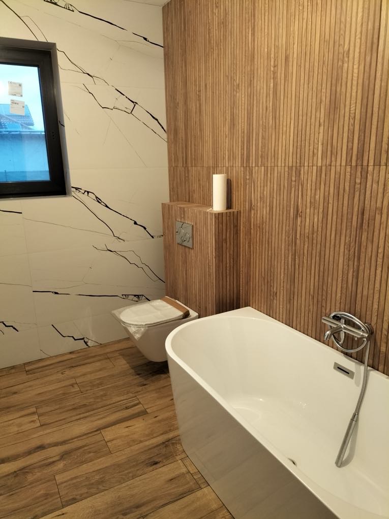 Nowoczesna łazienka z wanną, toaletą i drewnopodobnymi płytkami na ścianie oraz podłodze. Widoczne okno i minimalistyczna armatura.