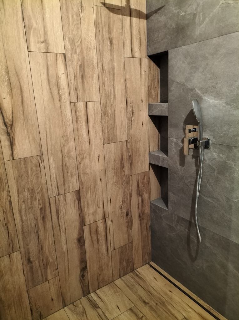 Nowoczesna kabina prysznicowa z płytkami imitującymi drewno na ścianie i podłodze, oraz szarą płytką na ścianie z wnękami i deszczownicą.