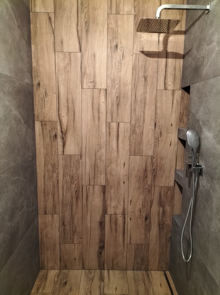 Kabina prysznicowa z drewnopodobnymi płytkami na ścianie i podłodze, szara ściana boczna, deszczownica i słuchawka prysznicowa.