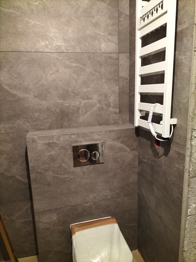 Szare, matowe płytki w łazience, chromowany przycisk spłuczki i biały grzejnik drabinkowy z kablem. Nowoczesne wykończenie.