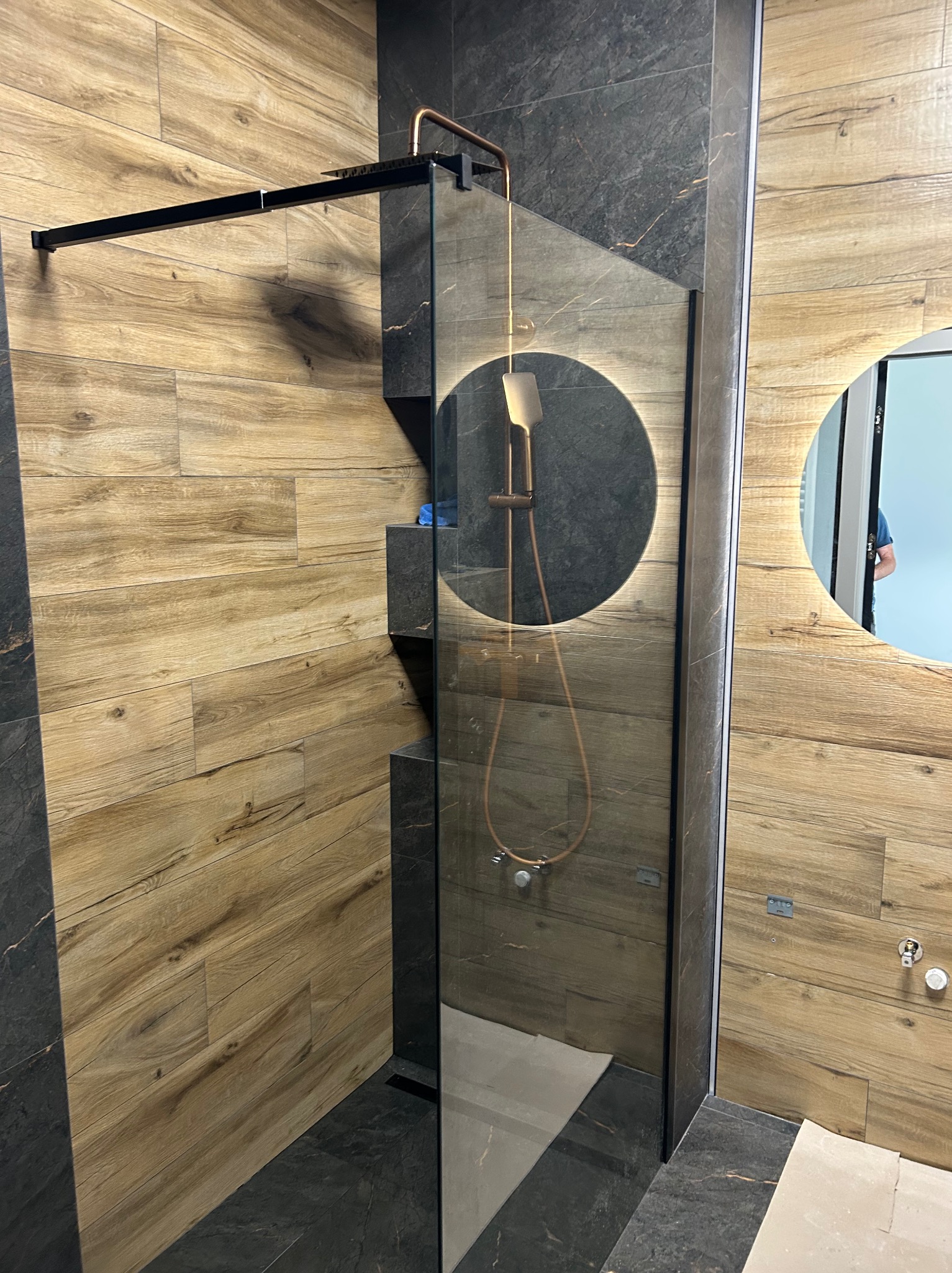 Nowoczesna kabina prysznicowa walk-in z deszczownicą i oświetleniem LED w okrągłym lustrze, wykończona płytkami imitującymi drewno i ciemnym kamieniem.