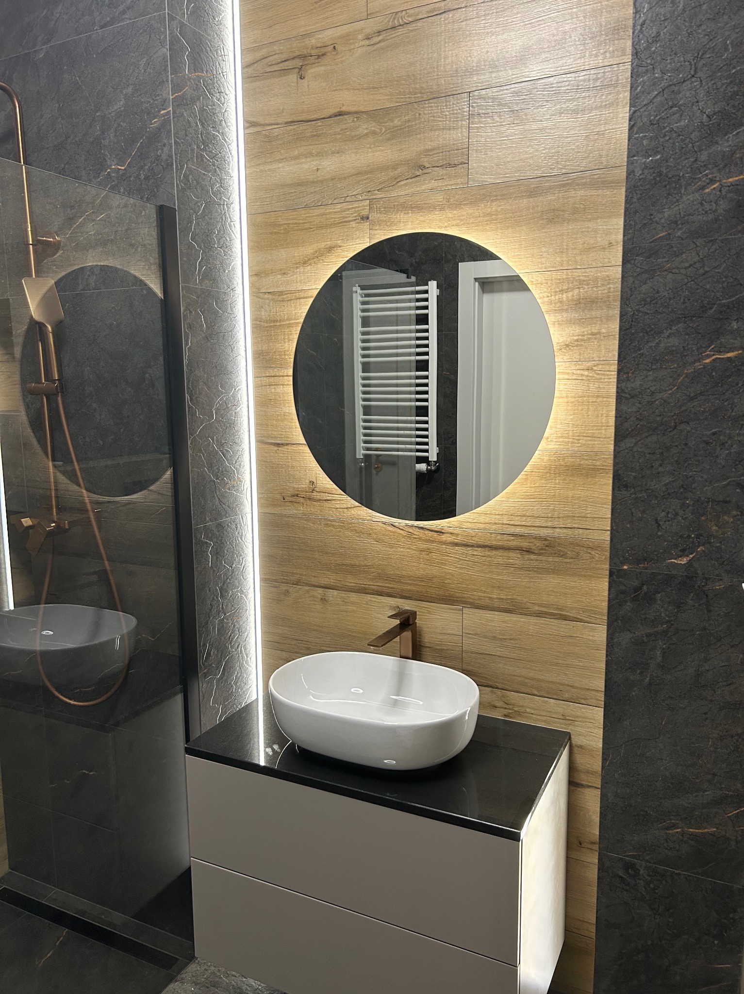 Nowoczesna łazienka z umywalką nablatową, okrągłym lustrem z podświetleniem LED, drewnopodobnymi i grafitowymi płytkami. Widoczny fragment kabiny prysznicowej.