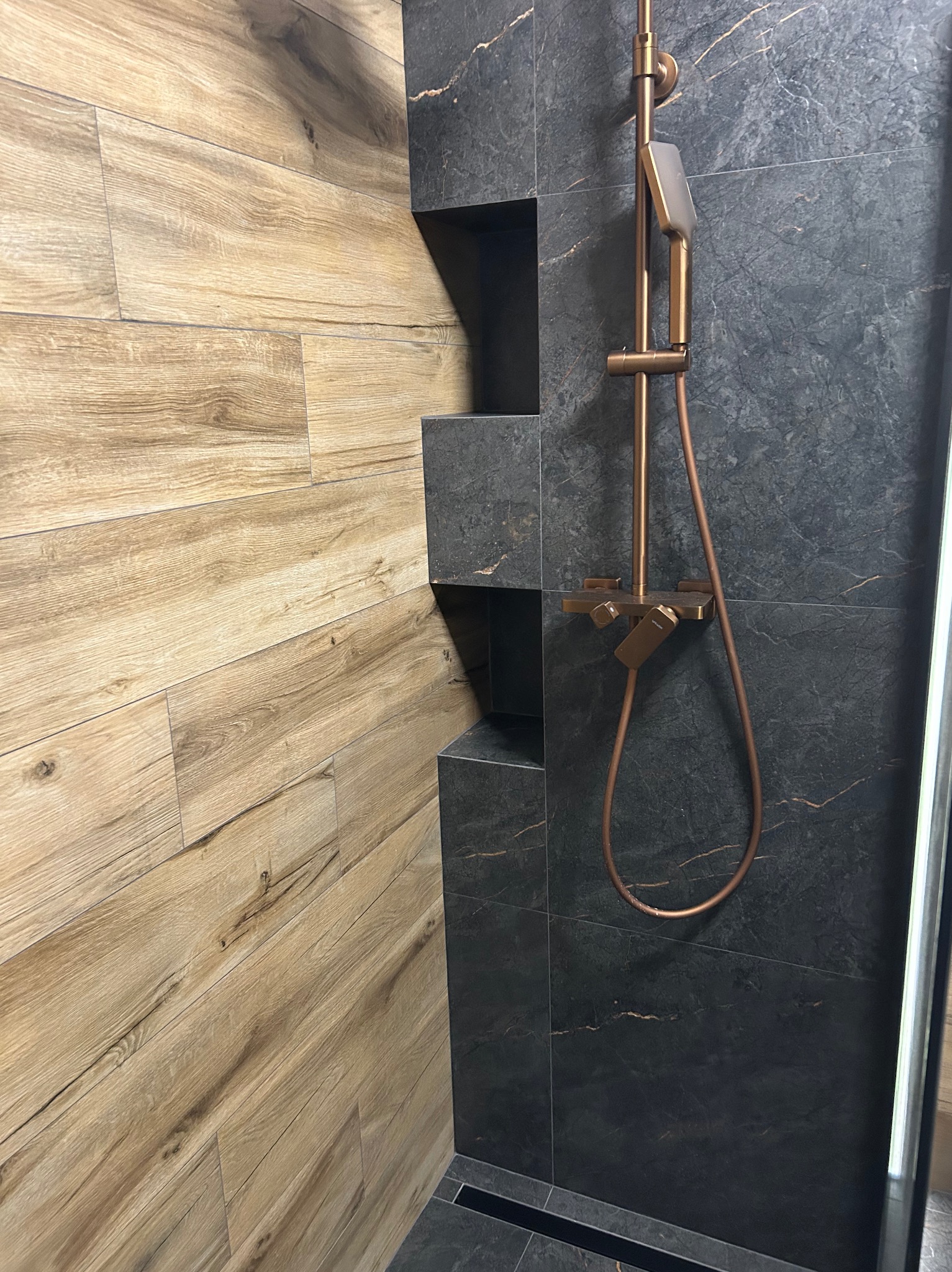 Nowoczesna kabina prysznicowa z panelami imitującymi drewno i ciemnymi płytkami imitującymi marmur. Miedziana armatura, wnęki w ścianie.