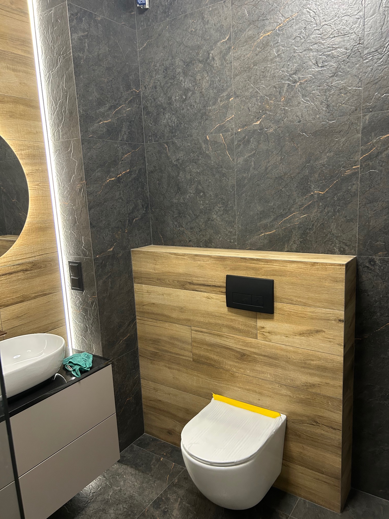 Nowoczesna łazienka z szarymi płytkami imitującymi kamień, drewnianą zabudową toalety i okrągłym lustrem z podświetleniem LED. Toaleta zabezpieczona folią.