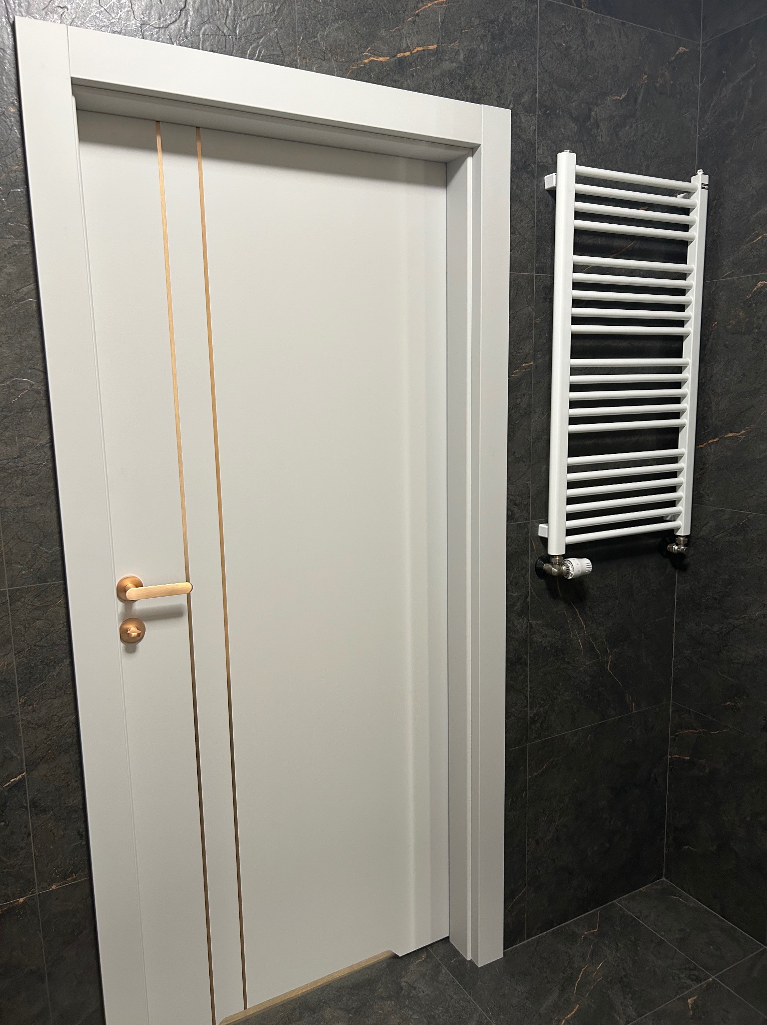 Białe drzwi z złotymi akcentami i grzejnik na tle ciemnej, marmurkowej ściany w łazience. Nowoczesny design, widoczne płytki podłogowe i minimalistyczne wykończenie.