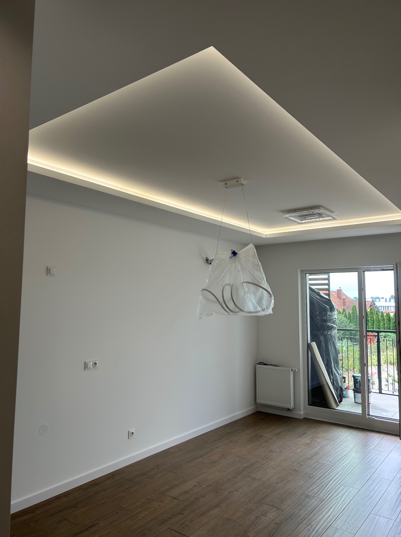 Nowoczesne wykończenie wnętrza: białe ściany, podwieszany sufit z oświetleniem LED, parkiet i zakryta lampa, w trakcie remontu, z widokiem na balkon.