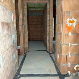 AZB Piotr Patoń - Wnętrze w trakcie budowy z widocznymi instalacjami elektrycznymi w peszlach prowadzonymi po podłodze, pomarańczowymi oznaczeniami punktów elektrycznych na ścianach z cegły dziurawki i wystającymi...