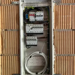 Rozdzielnica elektryczna w trakcie montażu w ścianie z cegły kratówki, widoczne przewody, bezpieczniki i elementy mocujące, ściana nieotynkowana.
