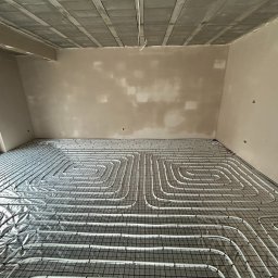 AZB Piotr Patoń - Białe rury ogrzewania podłogowego ułożone spiralnie na czarnej siatce w surowym wnętrzu z oknem i betonowym sufitem przed wylewką. Widoczne niedoskonałości ścian czekających na wykończenie.