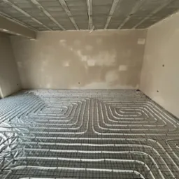 Białe rury ogrzewania podłogowego ułożone spiralnie na czarnej siatce w surowym wnętrzu z oknem i betonowym sufitem przed wylewką. Widoczne niedoskonałości ścian czekających na wykończenie.