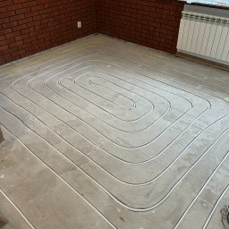 AZB Piotr Patoń - Ułożone rury ogrzewania podłogowego w jastrychu, widok z góry na spiralny układ, ściana z czerwonej cegły w tle, grzejnik pod oknem.