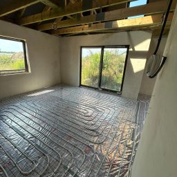 AZB Piotr Patoń - Rozłożone rury ogrzewania podłogowego na folii z siatką w surowym wnętrzu z widokiem na zewnątrz przez okna, drewniana konstrukcja dachu.