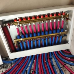 AZB Piotr Patoń - Rozdzielacz ogrzewania podłogowego z rurami w kolorach niebieskim i czerwonym, zamontowany w białej skrzynce, widoczne numery na zaworach, instalacja na siatce izolacyjnej.