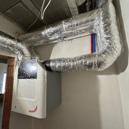 AZB Piotr Patoń - Centrala wentylacyjna Zehnder z izolowanymi przewodami wentylacyjnymi w surowym wnętrzu, widoczne rury instalacji wodnej i fragment niedokończonego muru.