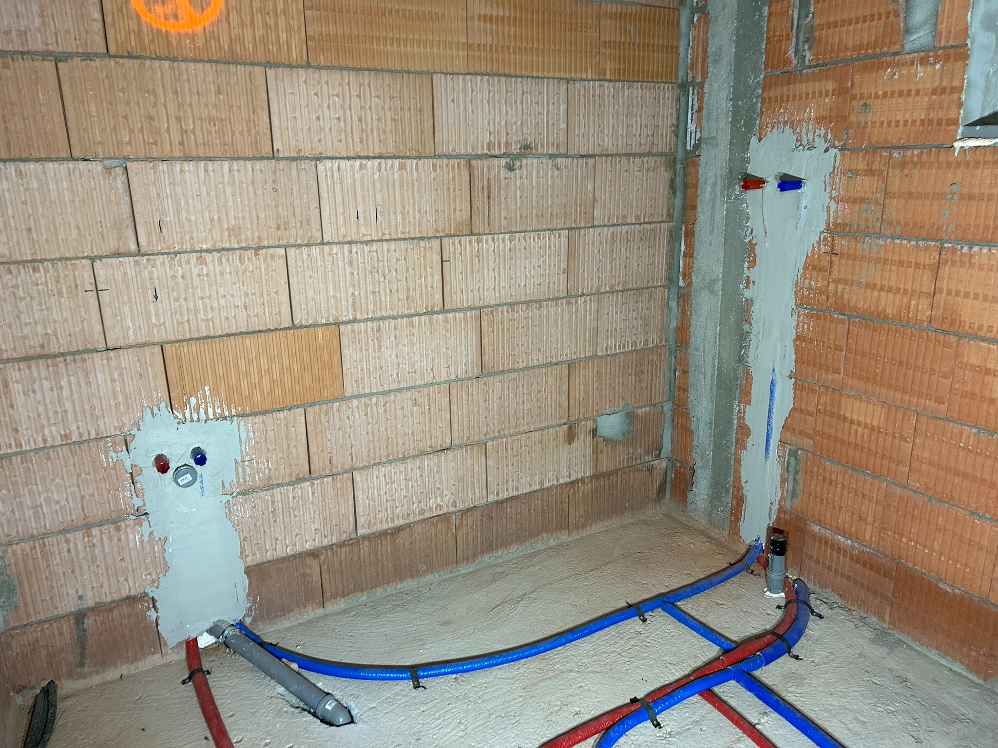 Surowy stan łazienki z widocznymi, częściowo zamurowanymi rurami instalacji wodnej w kolorach czerwonym i niebieskim, przymocowanymi do podłoża czarnymi opaskami zaciskowymi, oraz fragmentem pionu...