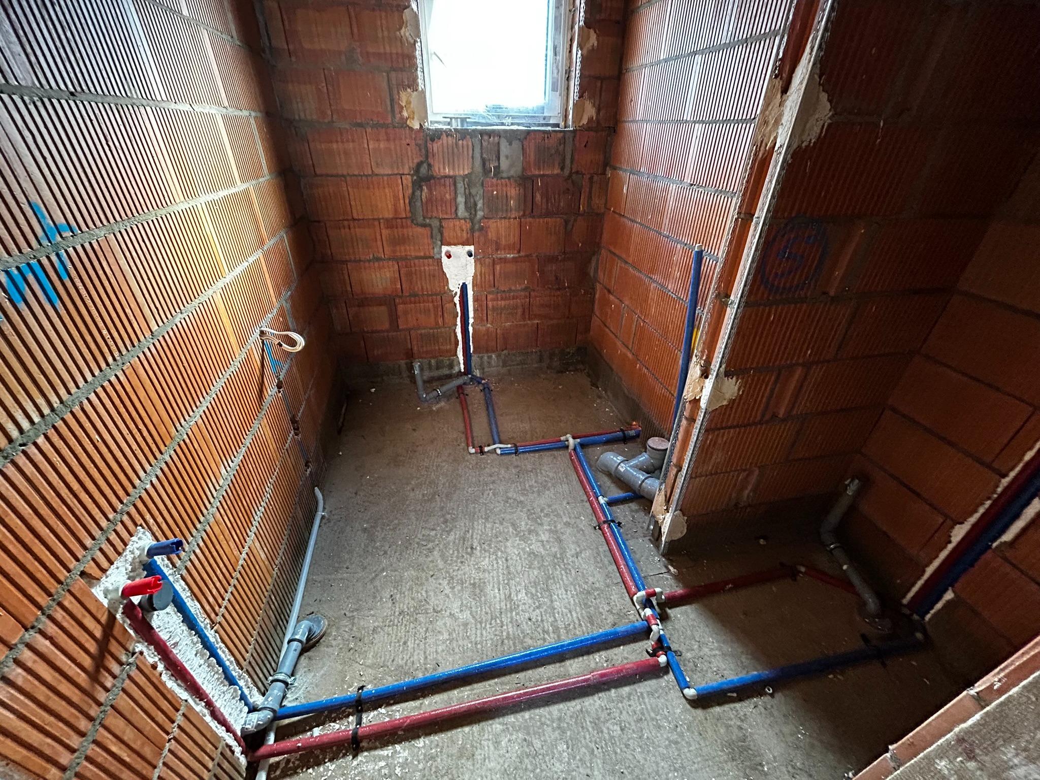 Surowe wnętrze łazienki w trakcie prac instalacyjnych, widoczne czerwone i niebieskie rury wodne prowadzone po podłodze i ścianach z cegły, przygotowane do podłączenia armatury.