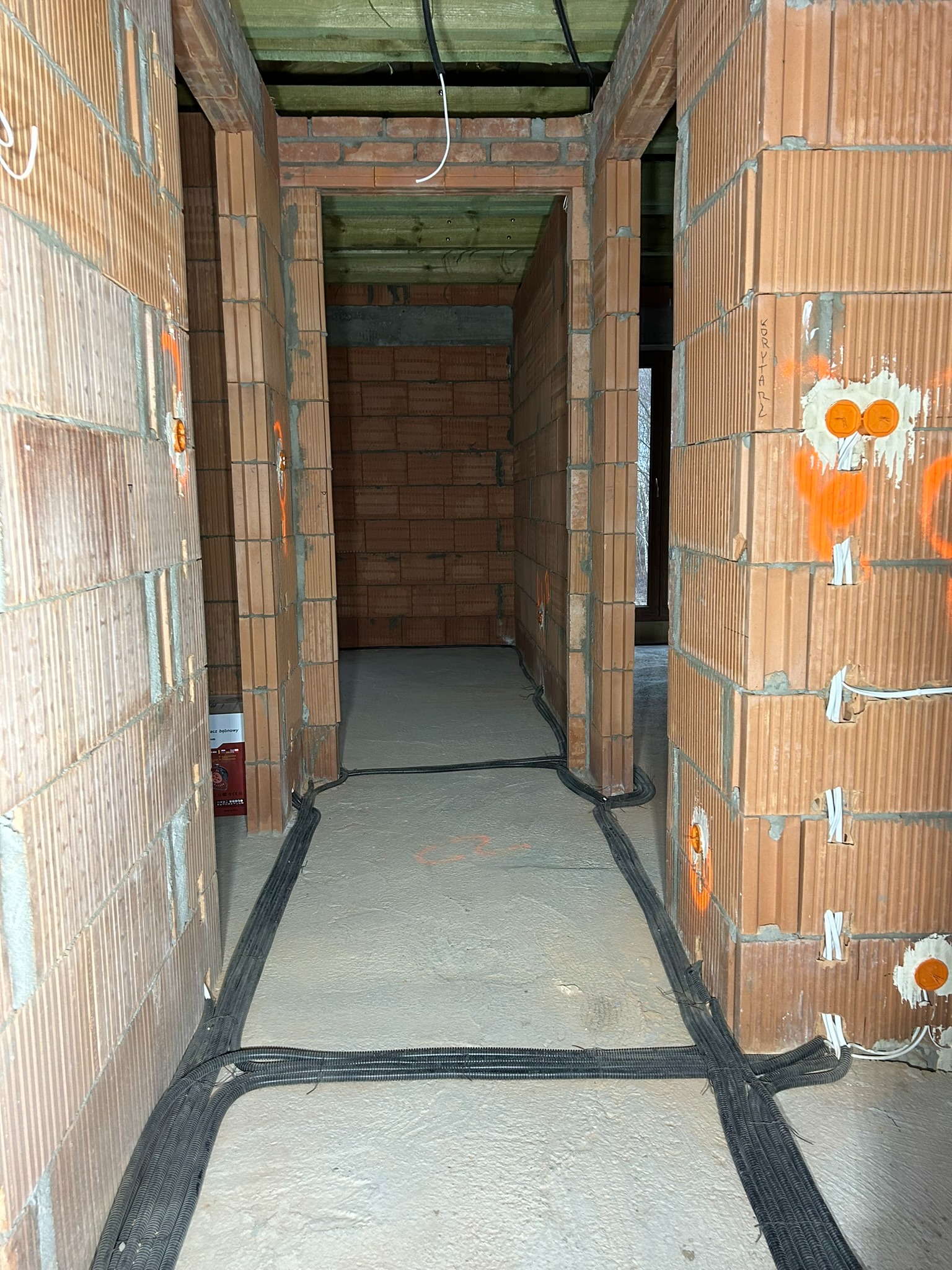 Wnętrze w trakcie budowy z widocznymi instalacjami elektrycznymi w peszlach prowadzonymi po podłodze, pomarańczowymi oznaczeniami punktów elektrycznych na ścianach z cegły dziurawki i wystającymi...