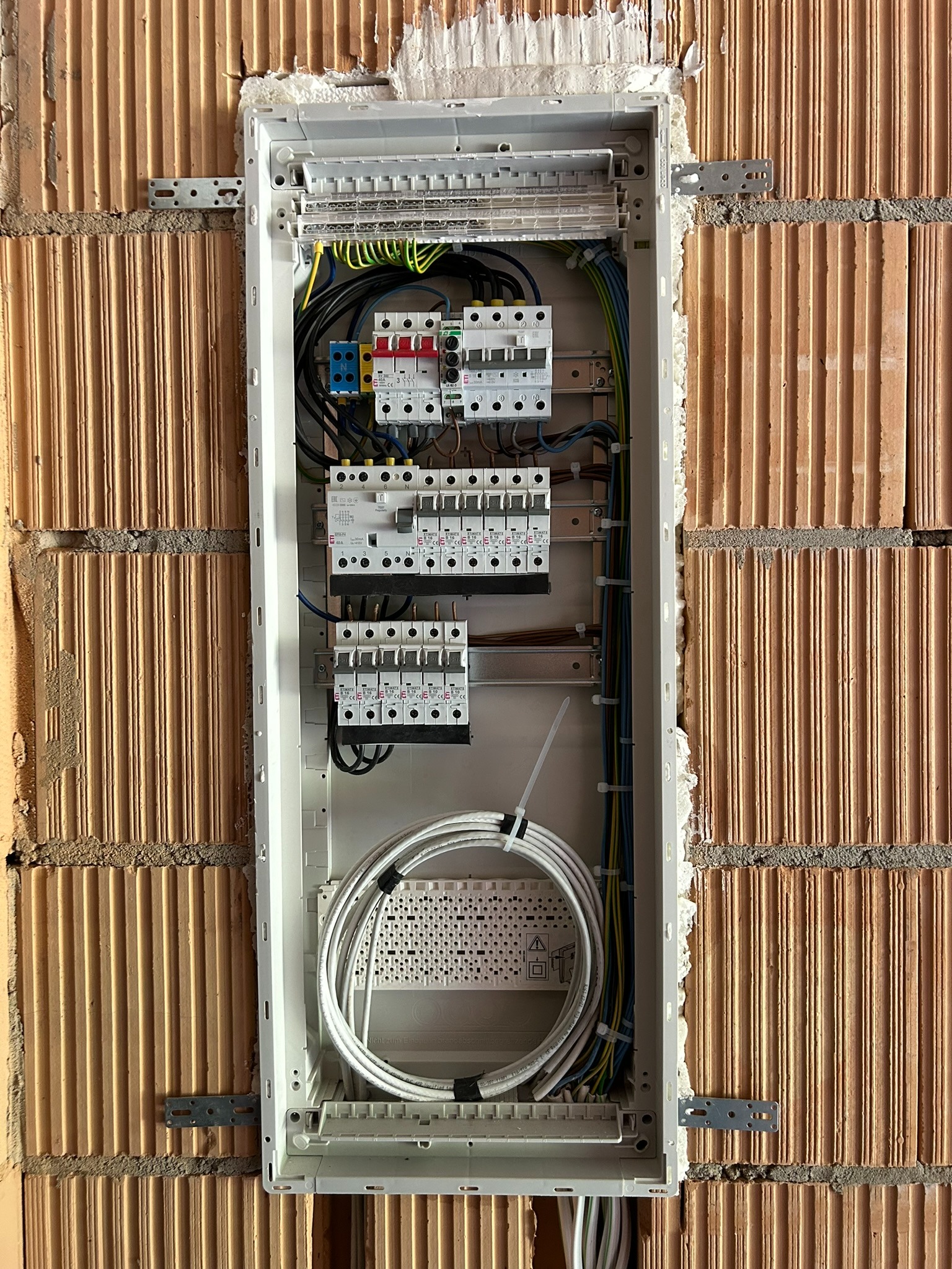 Rozdzielnica elektryczna w trakcie montażu w ścianie z cegły kratówki, widoczne przewody, bezpieczniki i elementy mocujące, ściana nieotynkowana.