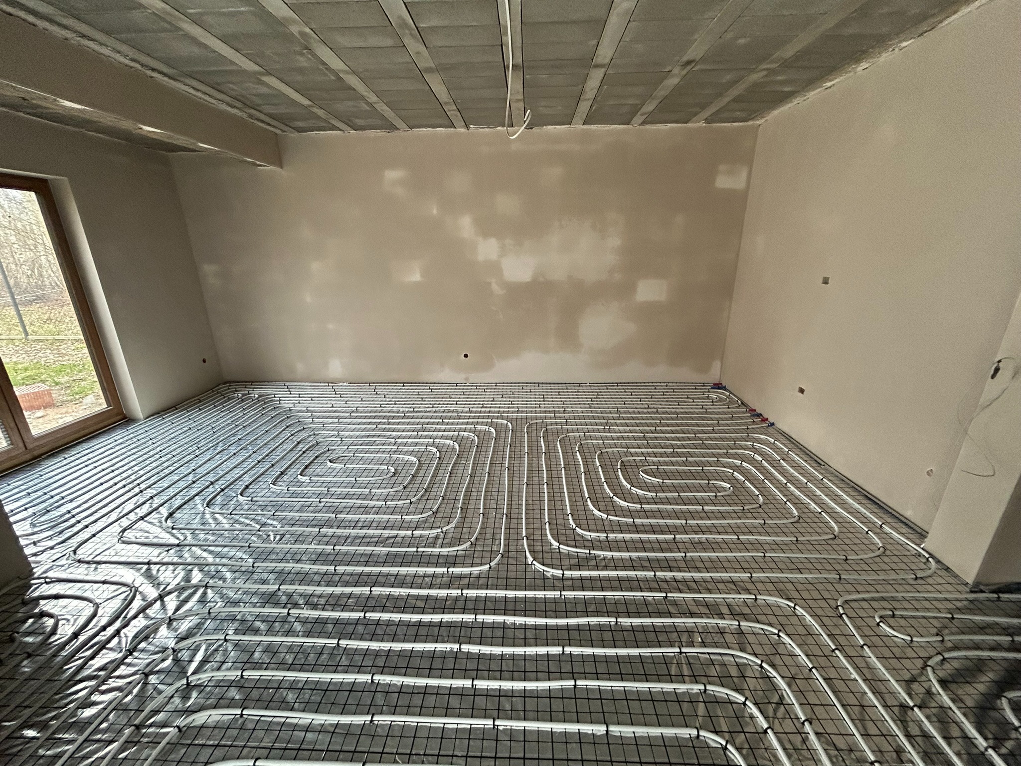Białe rury ogrzewania podłogowego ułożone spiralnie na czarnej siatce w surowym wnętrzu z oknem i betonowym sufitem przed wylewką. Widoczne niedoskonałości ścian czekających na wykończenie.