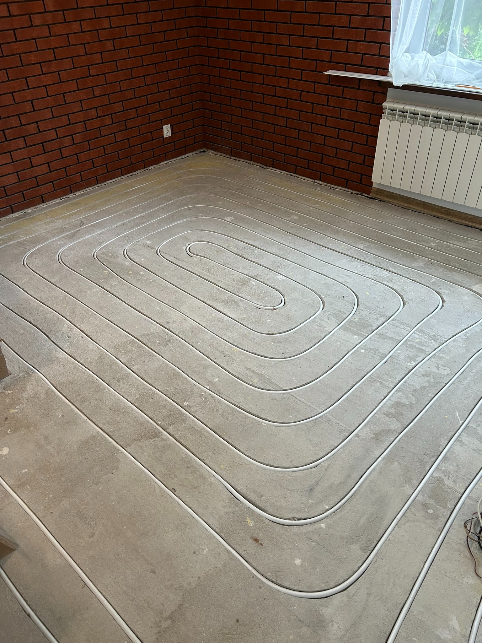 Ułożone rury ogrzewania podłogowego w jastrychu, widok z góry na spiralny układ, ściana z czerwonej cegły w tle, grzejnik pod oknem.