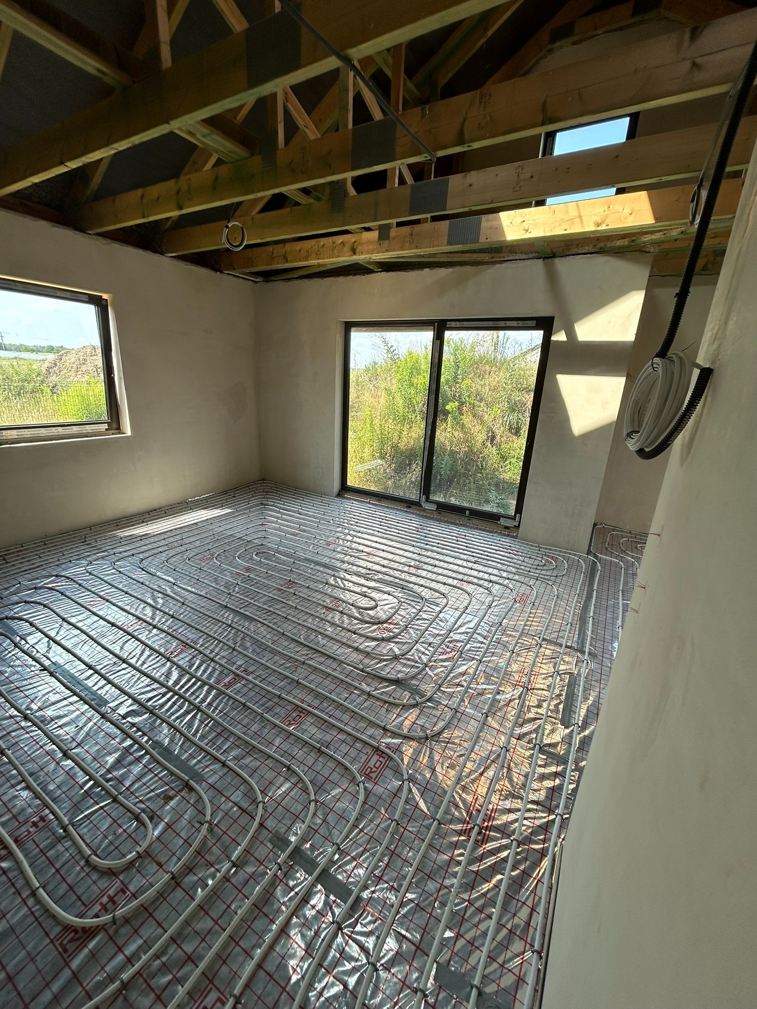 Rozłożone rury ogrzewania podłogowego na folii z siatką w surowym wnętrzu z widokiem na zewnątrz przez okna, drewniana konstrukcja dachu.