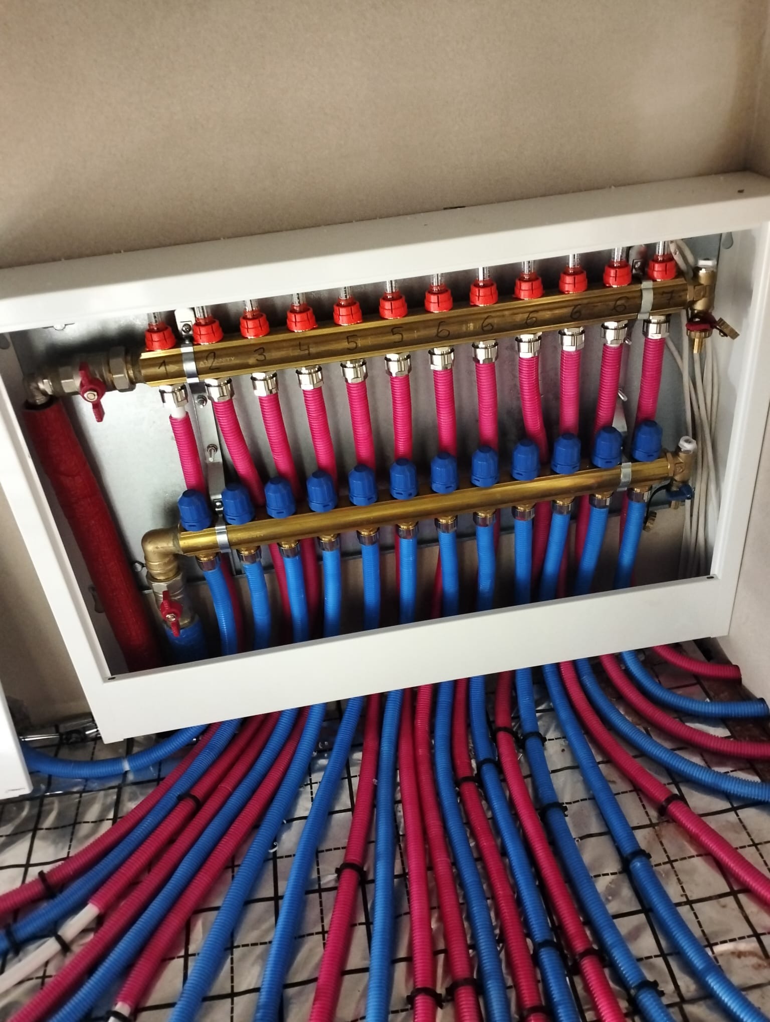 Rozdzielacz ogrzewania podłogowego z rurami w kolorach niebieskim i czerwonym, zamontowany w białej skrzynce, widoczne numery na zaworach, instalacja na siatce izolacyjnej.