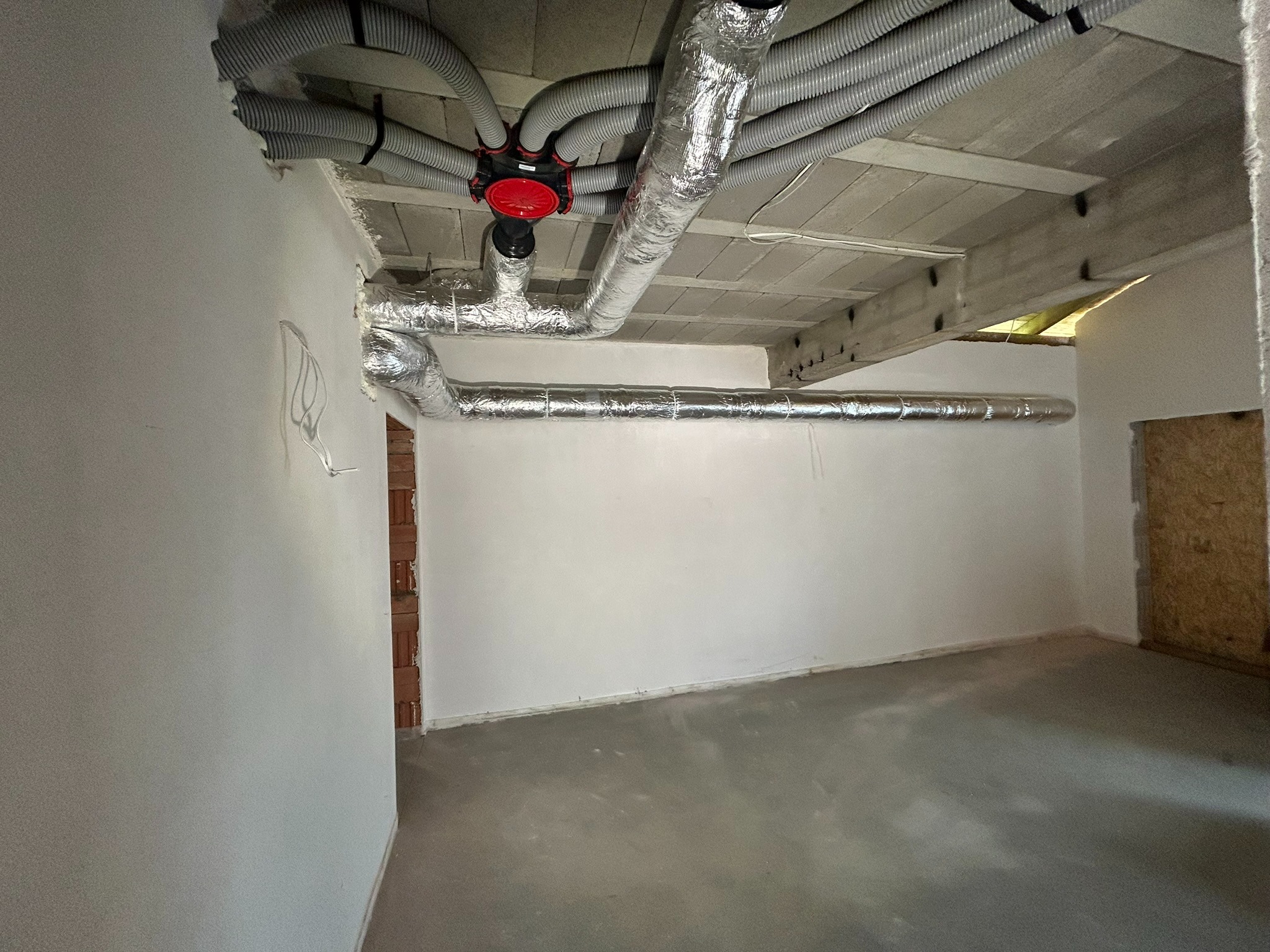 Surowy stan pomieszczenia z widoczną instalacją wentylacyjną: szare, elastyczne rury przymocowane do sufitu, rozchodzące się promieniście od czerwonego rozdzielacza, oraz srebrne, izolowane...
