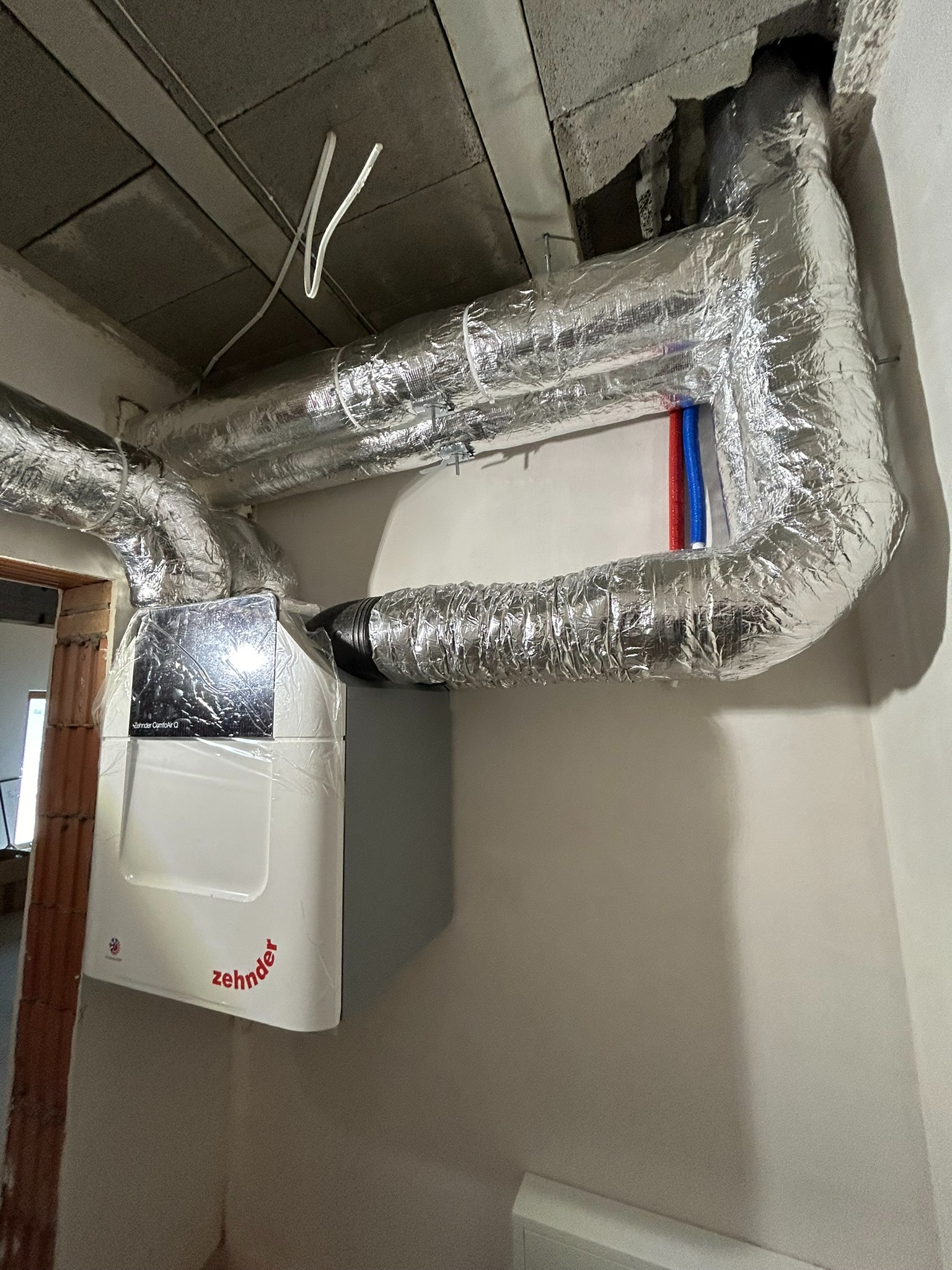 Centrala wentylacyjna Zehnder z izolowanymi przewodami wentylacyjnymi w surowym wnętrzu, widoczne rury instalacji wodnej i fragment niedokończonego muru.