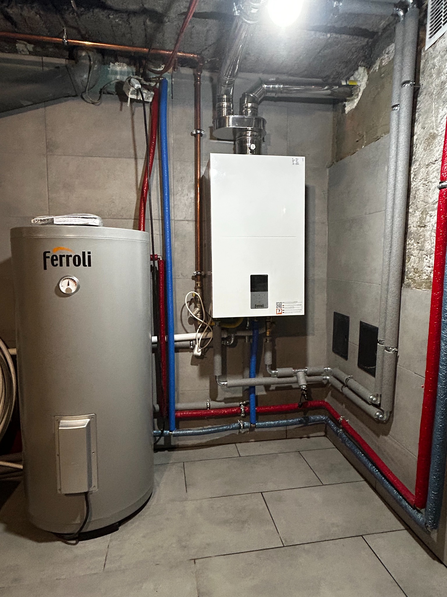 Kotłownia z zainstalowanym piecem gazowym Ferroli i zasobnikiem c.w.u., widoczne rury w izolacji termicznej w kolorach niebieskim i czerwonym, szare płytki na ścianach i podłodze.