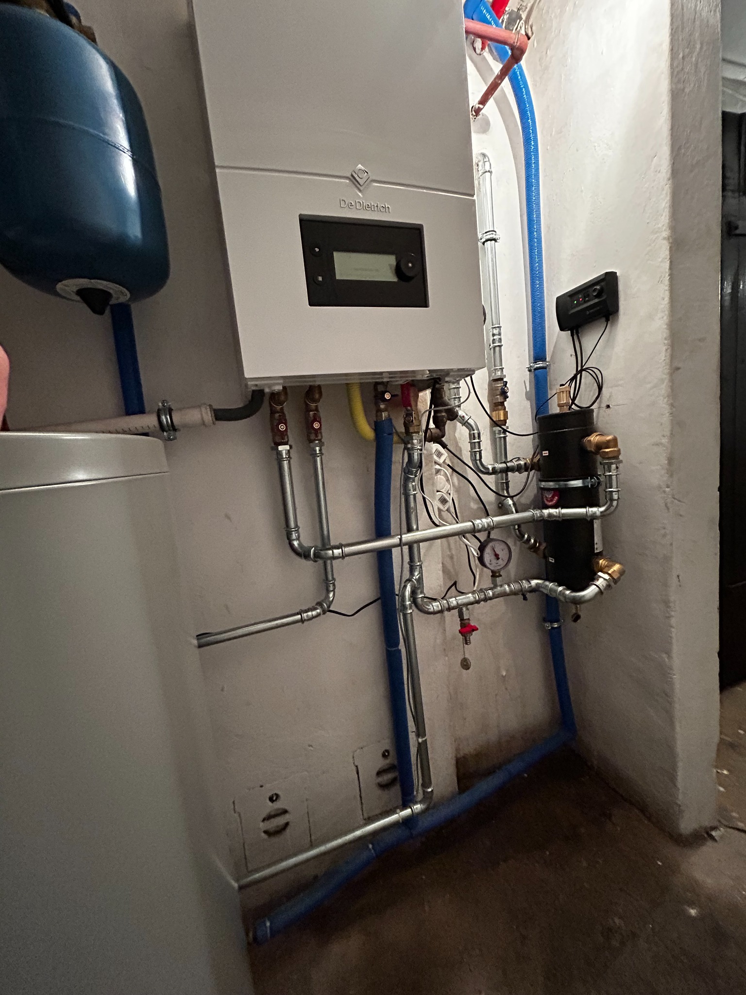 Nowoczesny piec gazowy De Dietrich zamontowany na ścianie, z widocznymi elementami instalacji, w tym rurami miedzianymi, zaworami oraz czarnym separatorem zanieczyszczeń, w pomieszczeniu o surowym...