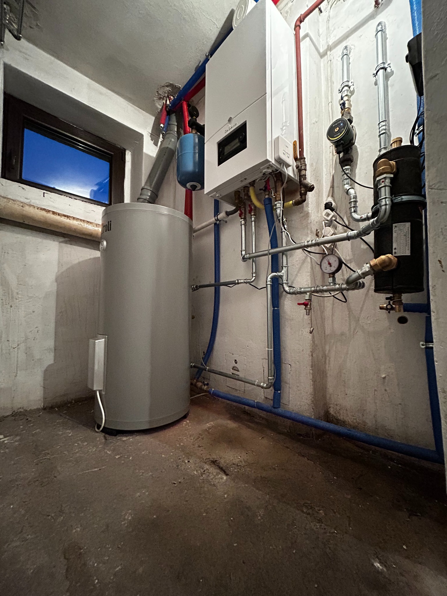 Nowoczesny piec gazowy wiszący obok szarego zasobnika ciepłej wody, rury w kolorze niebieskim i czerwonym, manometr i okno piwniczne z widokiem na nocne niebo w tle.