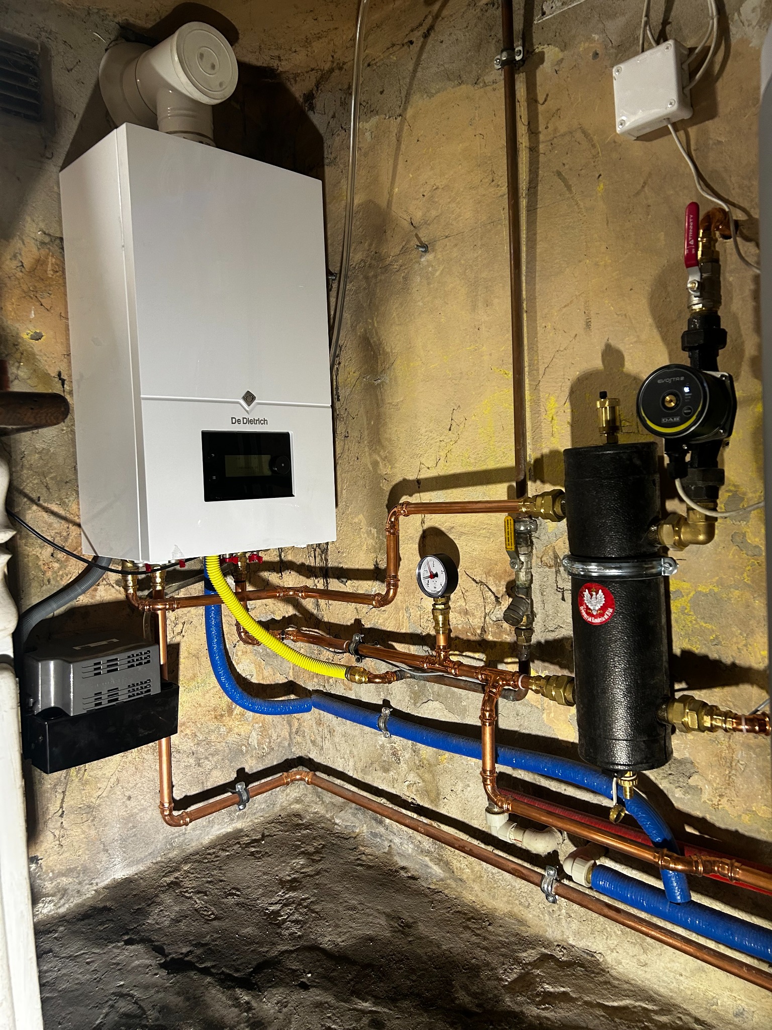 Zamontowany piec gazowy De Dietrich z podłączeniami miedzianymi i niebieskimi rurami PEX, widoczny również czarny zbiornik z naklejką, manometr i pompa obiegowa, na tle starej ściany.
