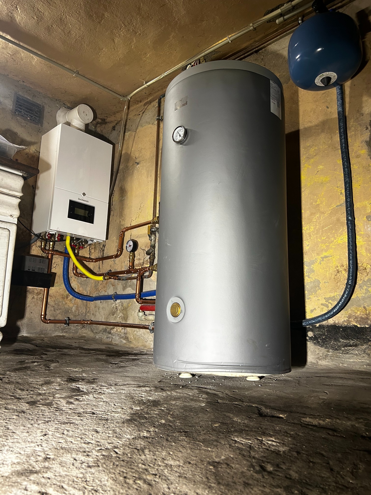 Nowoczesny piec gazowy De Dietrich z podłączeniami miedzianymi i zbiornikiem na wodę w piwnicy o surowych ścianach.