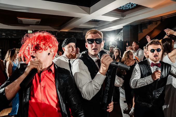 Zespół w trakcie występu na imprezie, wokalista w czerwonej peruce i skórzanej kurtce, drugi wokalista w pikselowych okularach trzyma mikrofon 'Super-Star', w tle bawiący się goście.