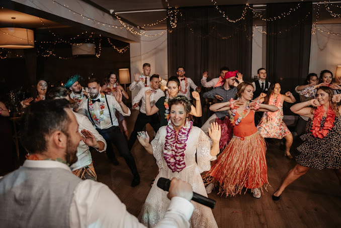 Grupa osób, w tym panna młoda z mikrofonem, tańcząca w hawajskich strojach na parkiecie podczas imprezy.
