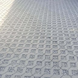 FIRMA OGÓLNOBUDOWLANA ERYK ŻYBURA - Szara, ażurowa kostka brukowa wypełniona drobnymi kamieniami, ułożona na posesji. Widoczny fragment trawnika i budynku w tle. Ujęcie z niskiej perspektywy.