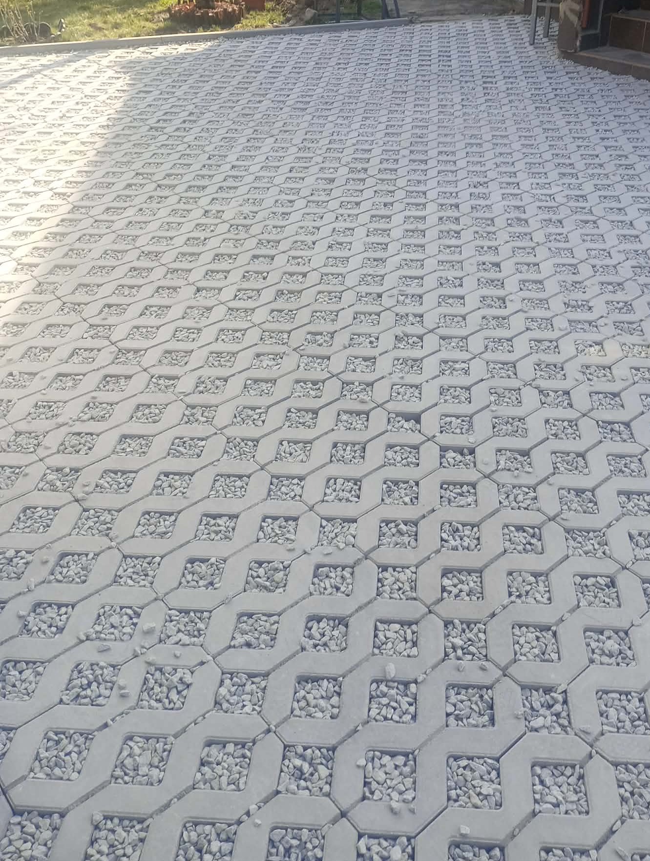 Szara, ażurowa kostka brukowa wypełniona drobnymi kamieniami, ułożona na posesji. Widoczny fragment trawnika i budynku w tle. Ujęcie z niskiej perspektywy.