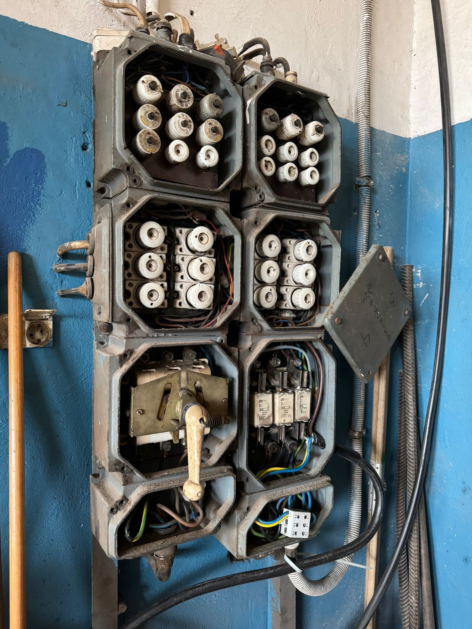 Stara, szara skrzynka bezpiecznikowa z ceramicznymi wkładkami, zamontowana na niebieskiej ścianie. Widoczne kable i elementy instalacji. Jeden z paneli jest otwarty.