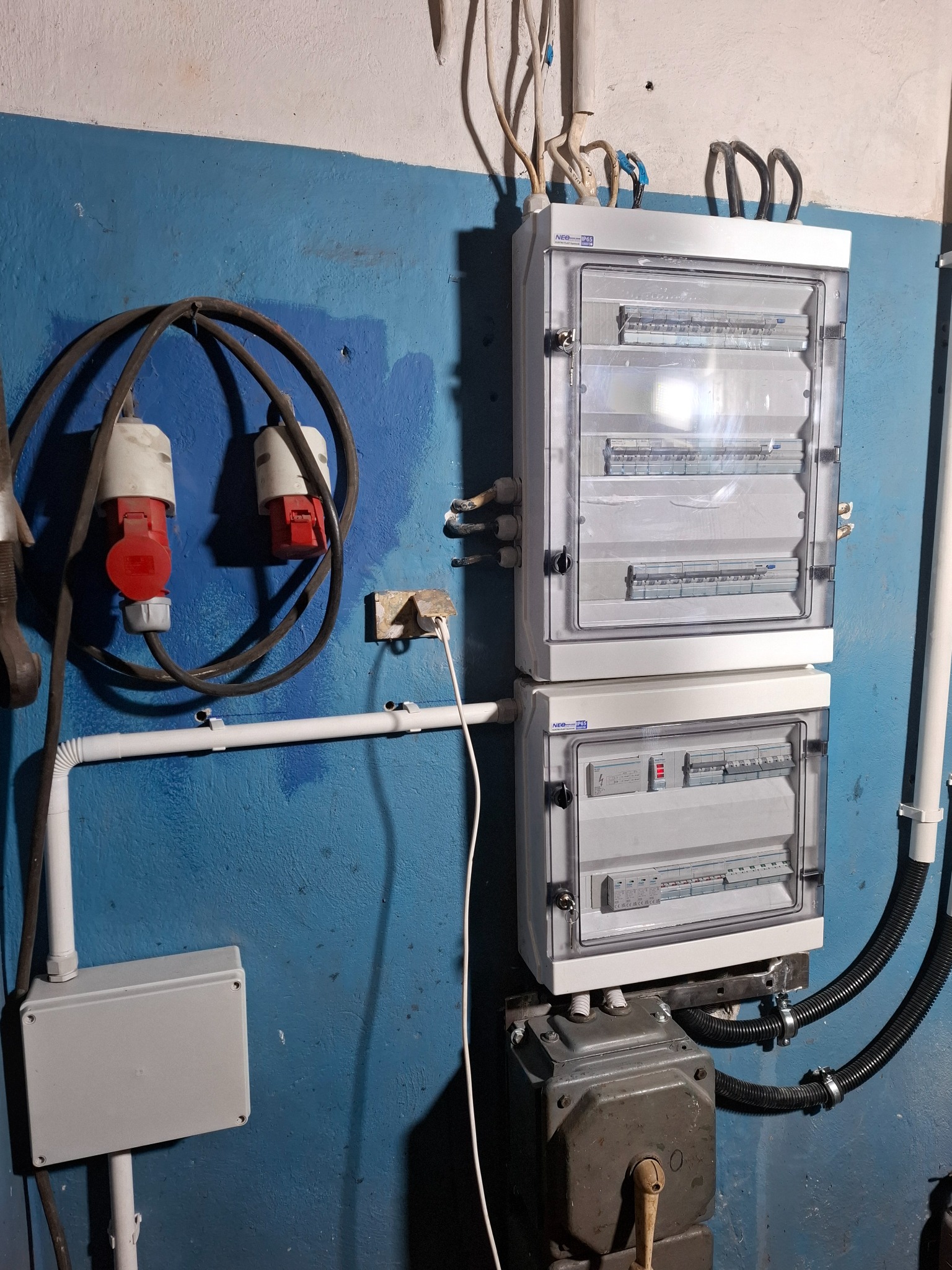 Dwa białe panele rozdzielcze z przezroczystymi drzwiczkami zamontowane na niebieskiej ścianie. Widoczne kable, rury i gniazda elektryczne.