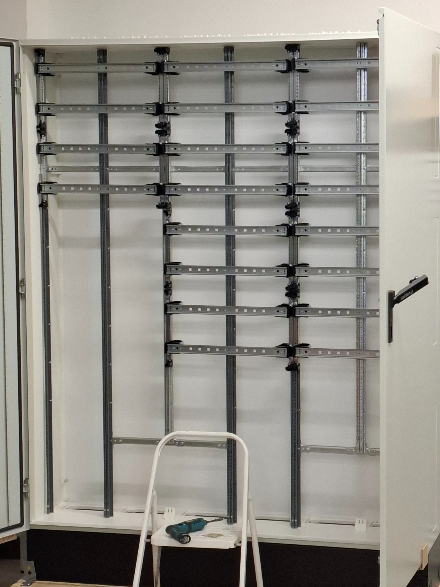 Wnętrze szafy elektrycznej z zamontowanymi szynami DIN i drabinkami kablowymi, widoczna drabina i wkrętarka na siedzisku. Białe tło, metalowe elementy.