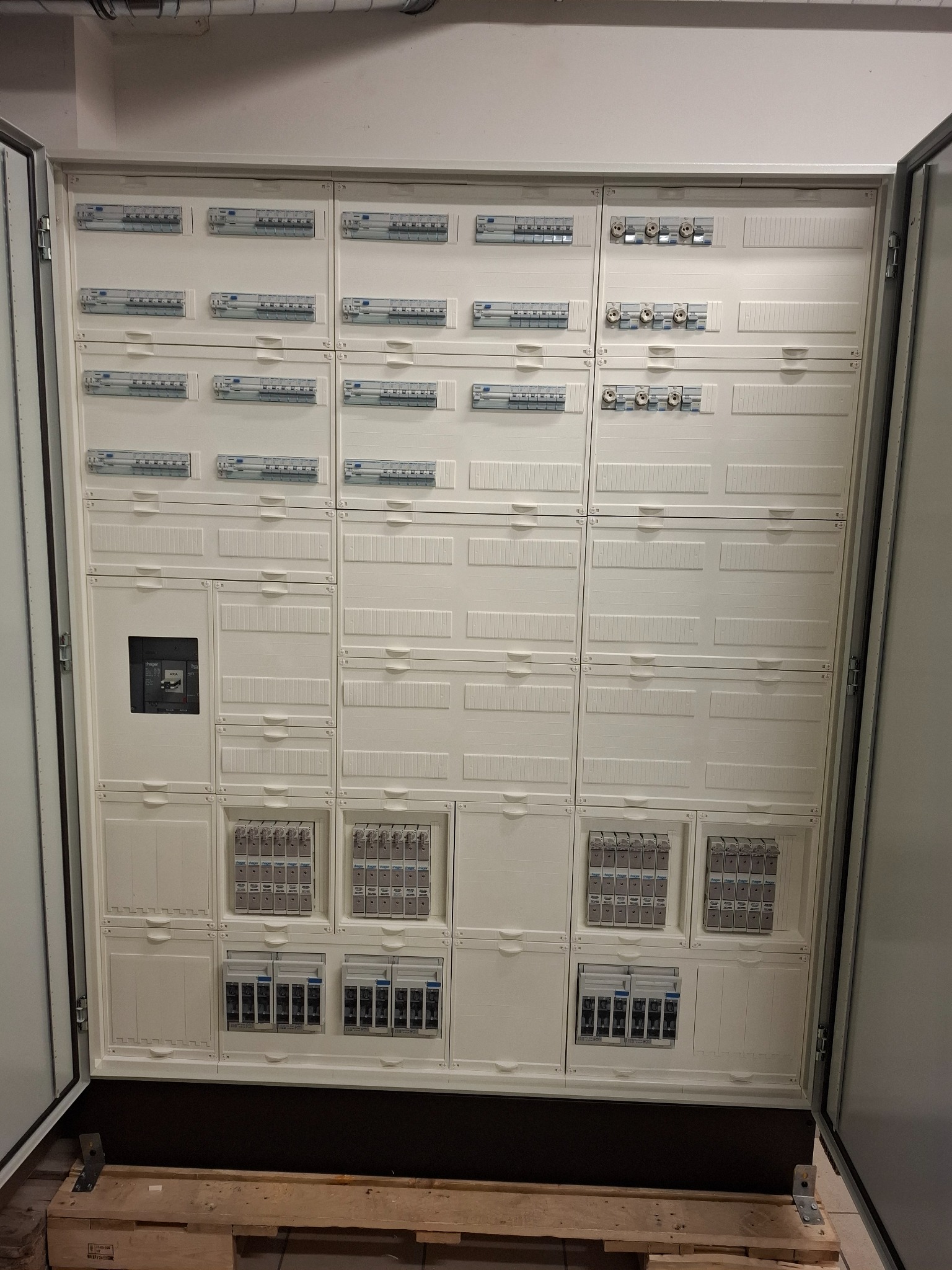 Otwarta, duża, biała szafa rozdzielcza z licznymi zabezpieczeniami i wyłącznikami, umieszczona na drewnianej palecie. Widoczne kable i rury na suficie.
