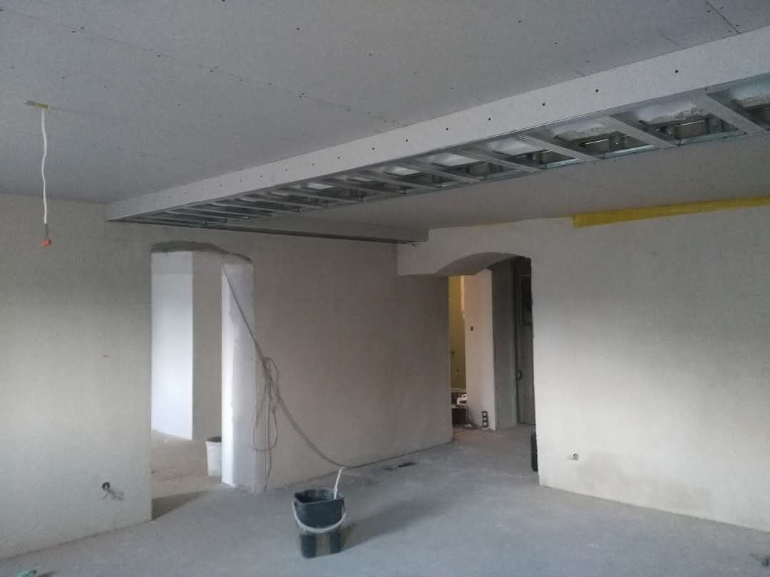 Wnętrze w trakcie prac wykończeniowych: surowy strop z odsłoniętą metalową konstrukcją, ściany przygotowane do gładziowania, widoczne wiadro i przewody.