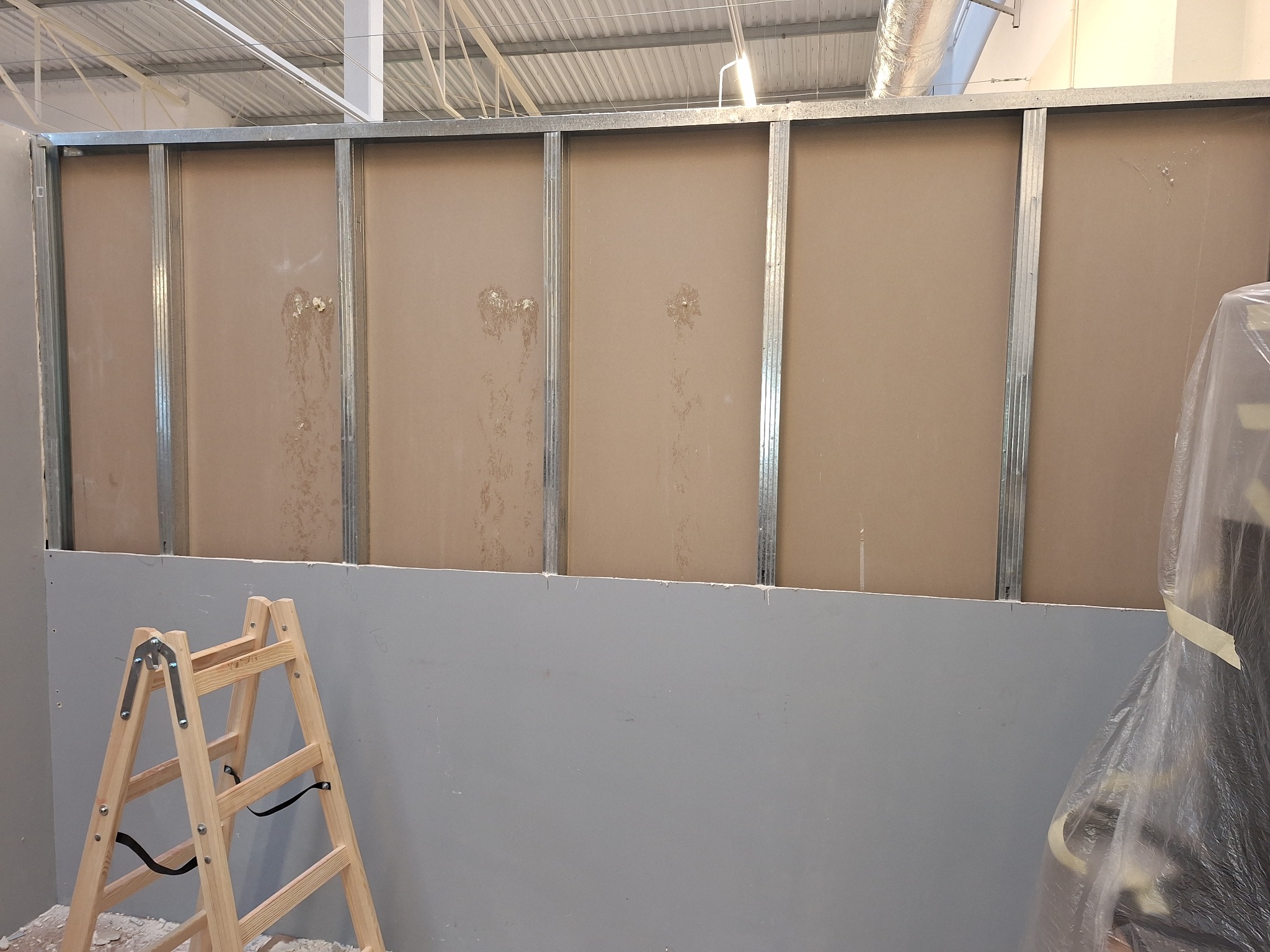Montaż stelaża z profili metalowych i płyt kartonowo-gipsowych, widoczna drewniana drabina i folia ochronna na elementach wyposażenia.
