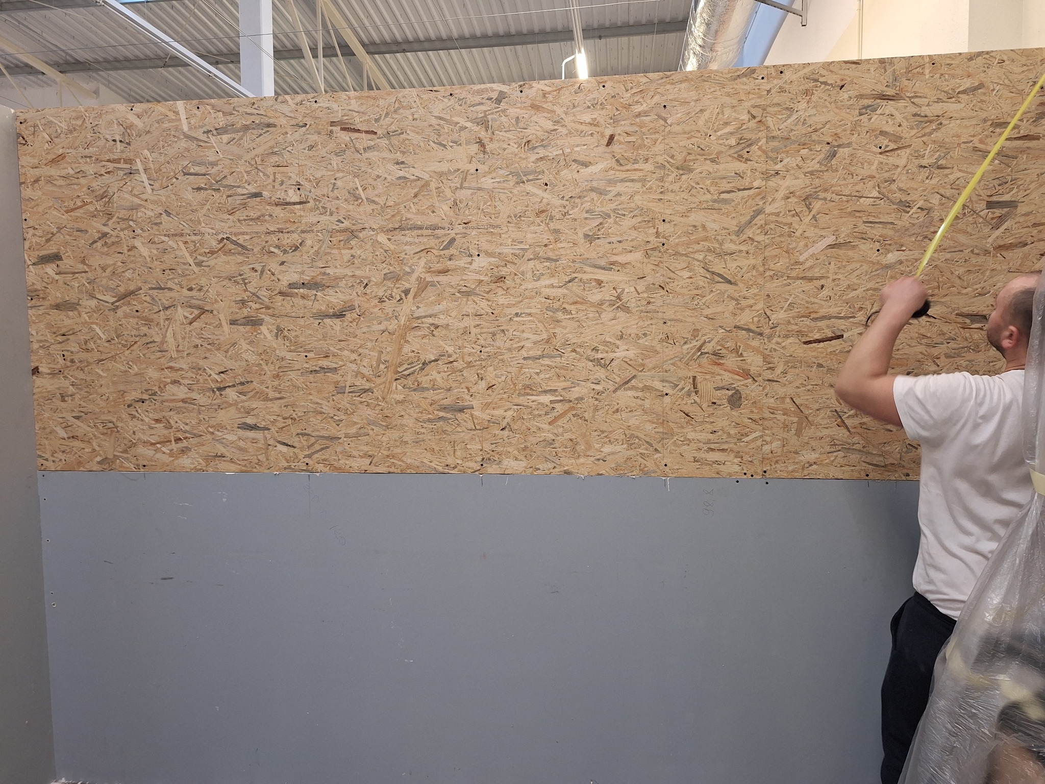 Mężczyzna mierzy żółtą miarką płytę OSB zamontowaną na szarej ścianie w pomieszczeniu z widoczną konstrukcją dachu.