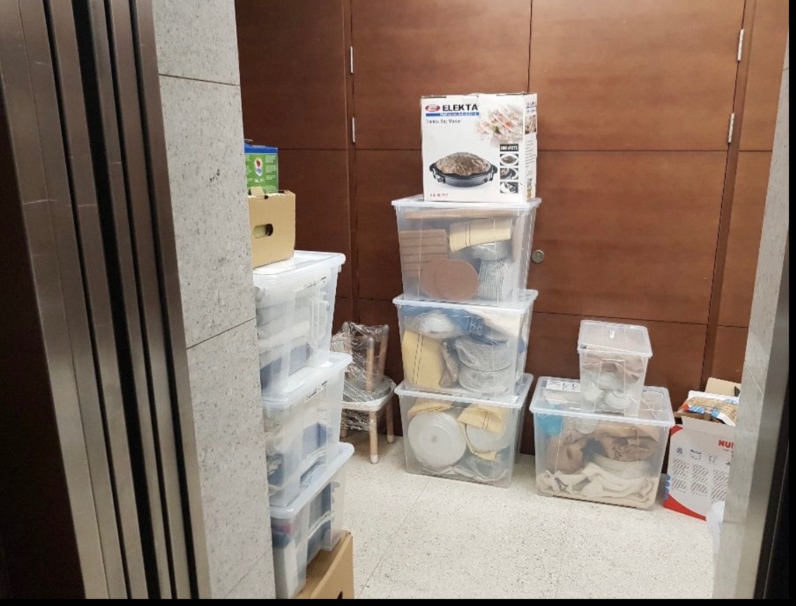 Stos plastikowych pojemników i kartonów z zawartością, przygotowanych do transportu podczas przeprowadzki. W tle widoczna ściana z brązowymi panelami.