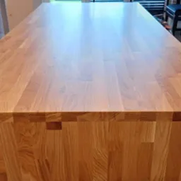 Długi, jasny blat z drewna klejonego, o wyraźnym usłojeniu, widoczny z góry, z fragmentem osoby odkurzającej krawędź, na tle mebli kuchennych.