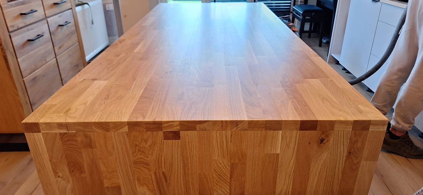 Długi, jasny blat z drewna klejonego, o wyraźnym usłojeniu, widoczny z góry, z fragmentem osoby odkurzającej krawędź, na tle mebli kuchennych.
